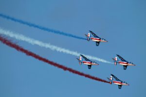 Four jets perform an aerobatic display leaving red, white, and blue smoke trails against a clear sky.